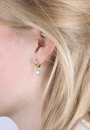 Gros plan sur l'oreille d'une femme blonde portant une petite boucle d'oreille en or avec un pendentif en pierre précieuse bleu clair.