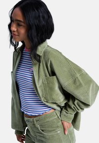 Πράσινο μπουφάν από κορδούρα με φερμουάρ και μπροστινή τσέπη, σε συνδυασμό με τοπ ριγέ σε μπλε και ροζ και παντελόνι κορδούρας σε ίδιο πράσινο χρώμα.