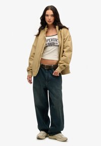 Veste zippée beige sur un crop top blanc à imprimé bleu, associée à un jean large bleu foncé et des baskets beiges.