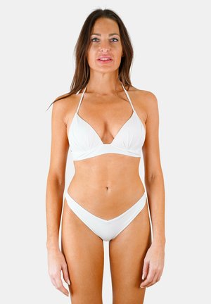 Femme portant un bikini deux pièces blanc avec un haut halter, debout devant un fond uni gris clair, face à la caméra.