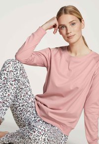 Light pink long-sleeve shirt paired with white pants featuring a floral pattern in pink and dark green, sitting pose on a neutral background.
