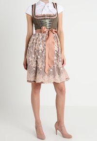 Mujer vistiendo un tradicional vestido dirndl verde y rosa con delantal de encaje y lazo de satén rosa, combinado con tacones altos beige.