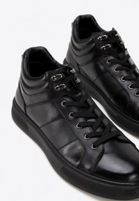 Black high-top sneakers made of smooth and textured leather, featuring round laces, metallic eyelets, and a rubber sole with patterned grip.