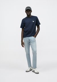 Μπλουζάκι σε στιλ navy με στρογγυλή λαιμόκοψη και μικρό λογότυπο, ανοιχτόχρωμα τζιν και λευκά αθλητικά παπούτσια με μαύριες ρίγες· μοντέλο που φοράει σκούρο καπέλο.