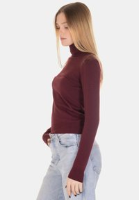 Maglione a collo alto color bordeaux in tessuto a maglia, design aderente, polsini e orlo a coste, indossato con jeans azzurri a vita alta.