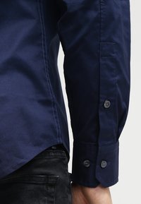 Mörkblå långärmad skjorta med manschett och två knappar, bärs med svarta denimjeans på en persons arm mot en neutral bakgrund.