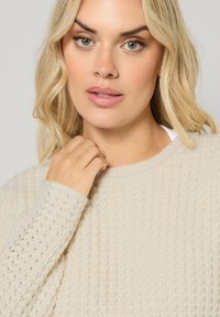 Pull en maille beige avec un motif texturé, encolure ronde et manches à motif. Modèle présentant le vêtement, avec des cheveux clairs encadrant le visage.