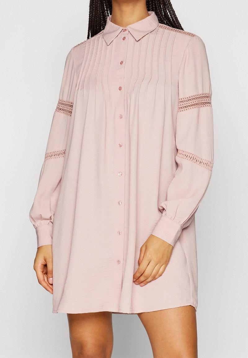 Helles rosa Hemdkleid mit langen Ärmeln, plissierter Textur und Häkelakzenten an den Ärmeln. Hergestellt aus einem weichen, leichten Material.