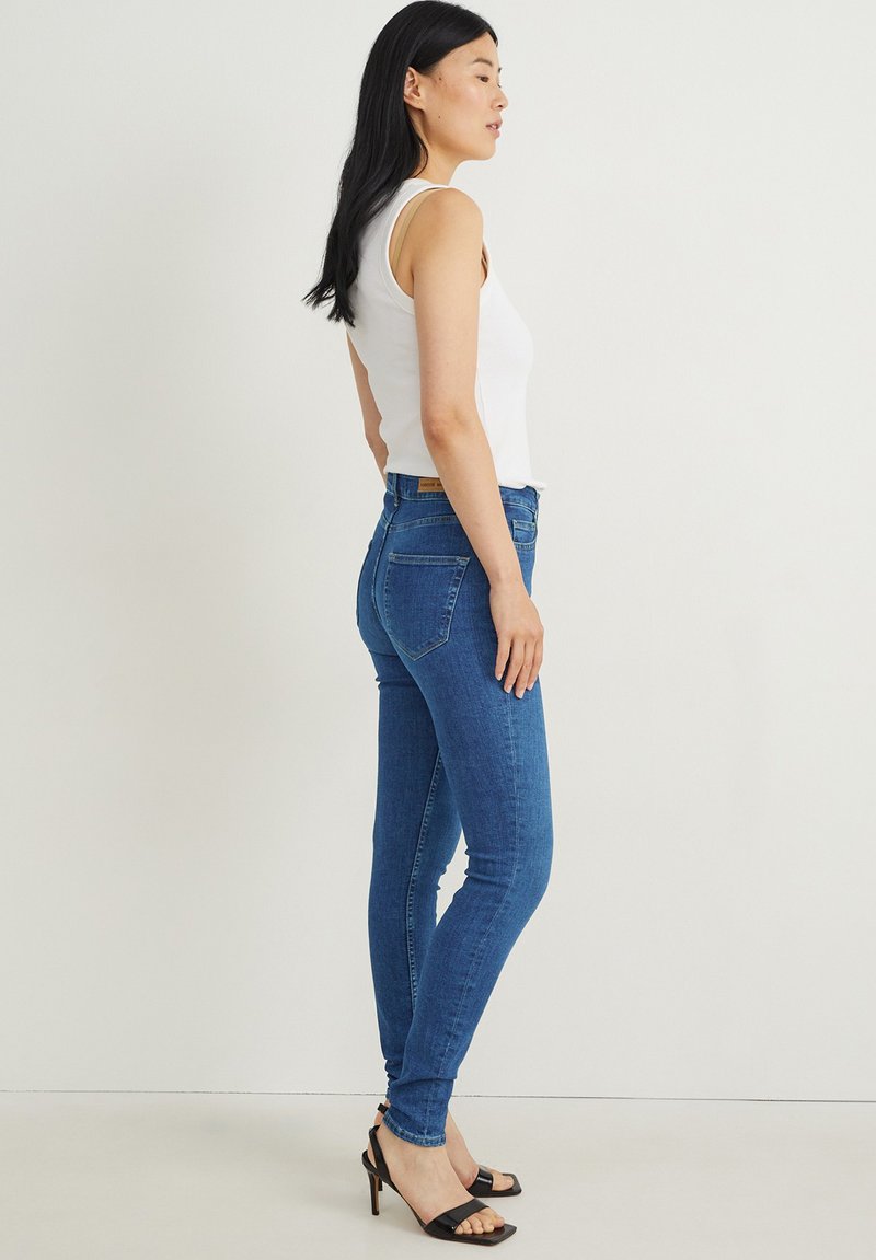 Schmale blaue Jeans aus Denim mit hoher Taille, fünf Taschen und dezentem Verblassen. Kombiniert mit einem weißen Tanktop und schwarzen Riemchenschuhen.