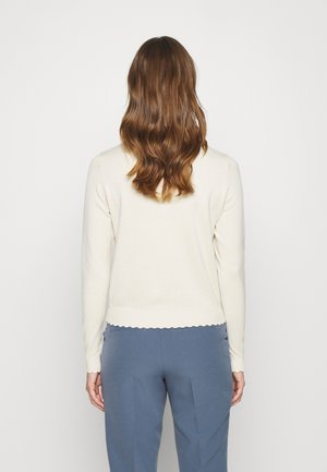Frau mit welligem braunem Haar, die ein weißes langärmliges Oberteil und blaue Hose trägt, steht mit dem Rücken zu einem schlichten hellen Hintergrund.