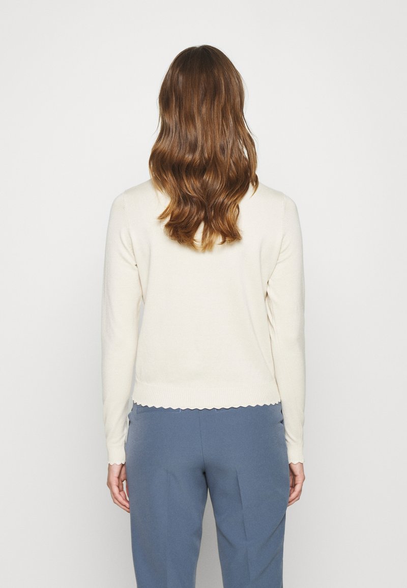 Femme aux cheveux bruns ondulés portant un haut blanc à manches longues et un pantalon bleu, debout de dos sur un fond clair uni.