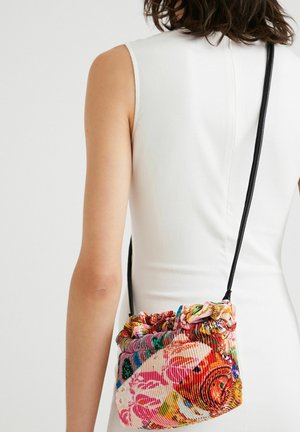 Frau in ärmellosem weißen Kleid trägt eine farbenfrohe Schultertasche mit Blumenmuster und schwarzem Riemen auf dem Rücken.