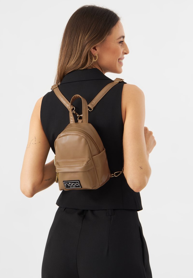 Zaino in finta pelle color beige dalla forma tondeggiante, con tasca zip e patch con il logo. Presenta spallacci regolabili e una maniglia superiore.