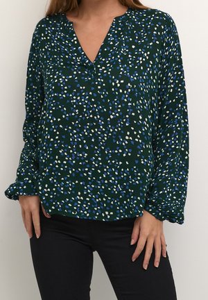 Kvinde iført mørkegrøn bluse med lange ærmer med blå og hvide pletter og sorte bukser, stående mod en ensfarvet baggrund.