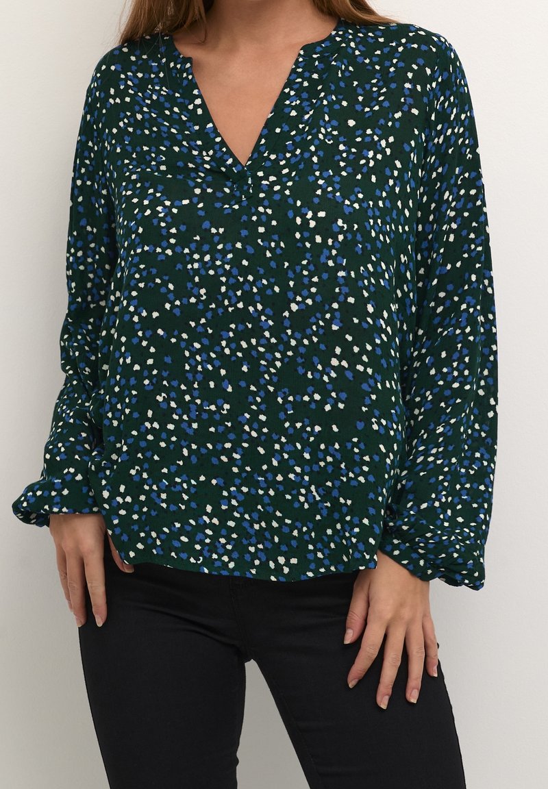 Femme portant un chemisier vert foncé à manches longues avec un motif tacheté bleu et blanc et un pantalon noir, debout devant un fond uni.