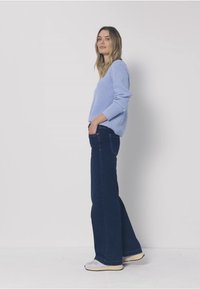 Pull en tricot bleu clair avec texture, associé à un jean évasé bleu foncé. Le mannequin porte des baskets grises. Fond simple pour un focus optimal.