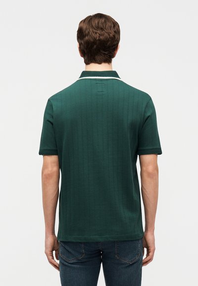 Άνδρας που φοράει σκούρα πράσινη κοντομάνικη πόλο μπλούζα με λευκή επένδυση στον γιακά και μπλε τζιν, φαίνεται από πίσω μπροστά σε απλό φόντο.