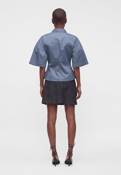 Personne portant une chemise bleue à manches courtes et une jupe noire, debout, dos tourné, avec des chaussures à talons noirs attachées aux chevilles.