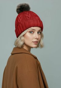 Red knit beanie with a thick ribbed cuff and cable pattern, topped with a fur pom-pom. Model wears a brown coat with a wide collar.