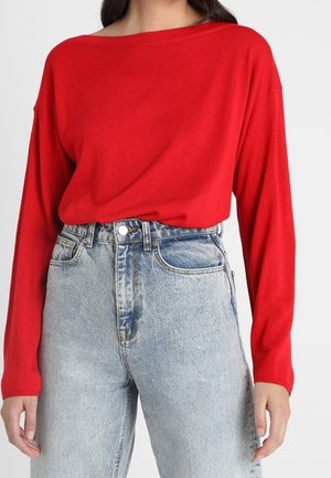 Frau trägt ein rotes Langarmoberteil, das in eine hoch taillierte hellblaue Jeans gesteckt ist, vor einem schlichten Hintergrund.