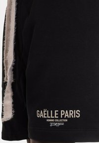 Pantaloni corti neri in cotone, con un accento sfilacciato di lato e testo stampato in beige. Il testo include "GAËLLE PARIS" e "HOMME COLLECTION."