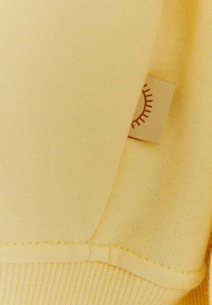 Žltá mikina so štruktúrovaným povrchom, s malou textilnou nášivkou so slnečným grafickým motívom. Švy sú starostlivo prešité a viditeľné.