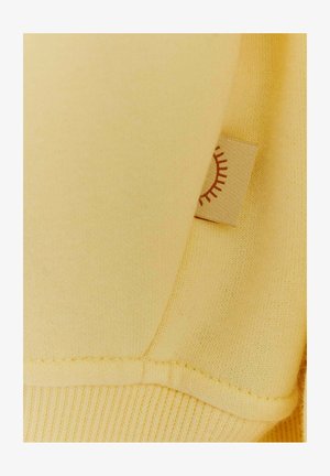 Żółta bluza z fakturowanym wykończeniem, z małą metką z tkaniny z graficznym wzorem słońca. Szwy są starannie przeszyte i widoczne.