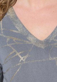 Pull V en gris avec des motifs abstraits dorés métalliques et un tricot texturé. Le col est orné d'une bordure brillante et pailletée.