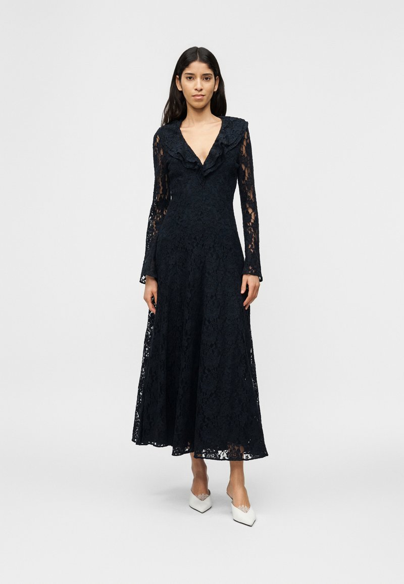 Γυναίκα που φοράει ένα μακρύ μαύρο δαντελένιο φόρεμα με βολάν στον V-λαιμό και μακριά μανίκια, συνδυασμένο με λευκά μυτερά τακούνια, στέκεται μπροστά σε απλό φόντο.