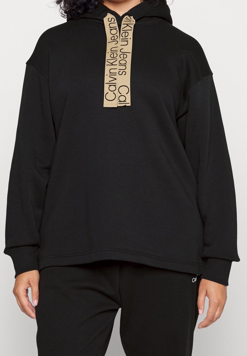 Hoodie noir avec une grande poche avant, doté d'un cordon beige portant la marque "Calvin Klein Jeans". Tissu doux et coupe ample.