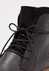 Stivaletto in pelle nera con una fascia in camoscio alla caviglia, caratterizzato da lacci rotondi e una finitura liscia. Include perforazioni lungo il lato per la ventilazione.