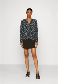 Mulher em pé, vestindo uma blusa de manga longa com decote em V em florais escuros, uma saia mini assimétrica preta e mules de salto aberto pretos, num fundo neutro.