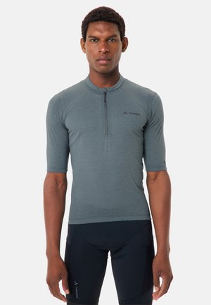 Man draagt een grijs wielershirt met halve ritssluiting en zwarte gewatteerde wielershorts, staande voor een effen lichte achtergrond en kijkt recht vooruit.