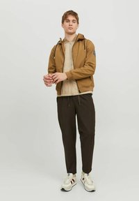 Veste brune zippée avec capuche, doublure beige matelassée, cordons de serrage noirs, associée à un pantalon sombre et des baskets blanches avec des accents rouges.