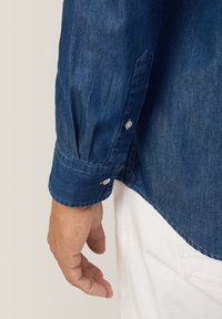 Camicia di denim blu scuro, con polsini button-down e cuciture bianche, orlo curvo, abbinata a un tessuto bianco visibile alla vita.