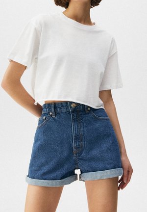 Person iført en helt hvid cropped t-shirt og højtaljede blå denimshorts med opkørte kant, stående med den ene hånd på hoften.