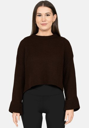 Vrouw met lang bruin haar, gekleed in een donkerbruine grofgebreide trui en zwarte aansluitende broek, staand tegen een witte achtergrond.