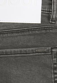 Jeans in denim nero con un'etichetta in pelle bianca a vista con branding embossato e un piccolo cartellino metallico con la scritta "HUGO" sulla tasca.