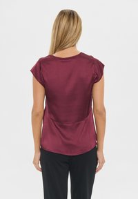 Burgundy kortärmad blus med en slät, glänsande textur. Har en rund halsringning och en böjd nederkant, tillsammans med svarta byxor.