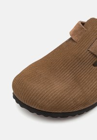 Hnedé kordurové nazúvacie topánky s čiernou drážkovanou podrážkou a pásikom cez vrch, zobrazené na bielom pozadí.