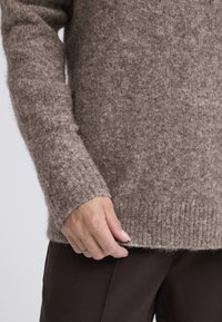 Rjav pulover iz pletenine z teksturirano, puhasto površino, rebrastimi manšetami in rahlo preveliko prileganjem, kombiniran s temno rjavimi hlačami.