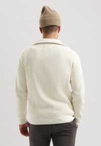 Pulover crem cu striații și guler, asortat cu pantaloni închiși. Modelul poartă o căciulă tricotată de culoare maro deschis.