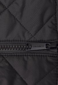 Tissu matelassé noir avec une fermeture éclair horizontale dotée d'une languette texturée. La tirette de la fermeture éclair est marquée "carhartt".