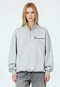 Sweat-shirt gris avec un col demi-fermeture éclair, arborant un texte noir en cursive « Amour » sur la poitrine gauche. Texture douce et coupe décontractée.