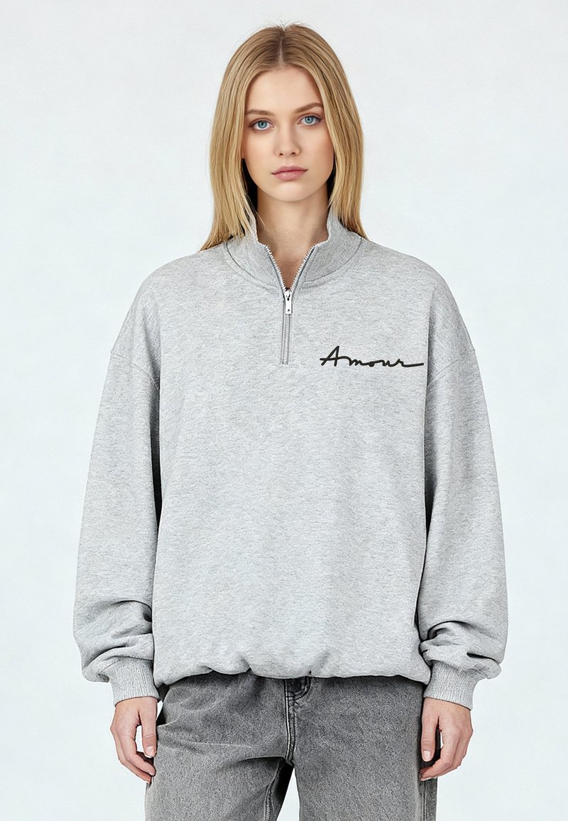 Sweat-shirt gris avec un col demi-fermeture éclair, arborant un texte noir en cursive « Amour » sur la poitrine gauche. Texture douce et coupe décontractée.