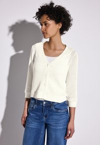 Cardigan tricoté crème clair avec un décolleté en V, fermeture à boutons et manches 3/4, associé à un jean bleu. Le tissu texturé ajoute un intérêt visuel.