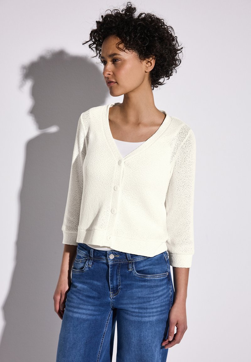 Cardigan tricoté crème clair avec un décolleté en V, fermeture à boutons et manches 3/4, associé à un jean bleu. Le tissu texturé ajoute un intérêt visuel.