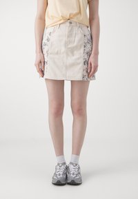 Lys beige denimskjørt med blomsterpaneler på siden, knappelukking og rett passform. Brukt med grå sneakers og hvite sokker.