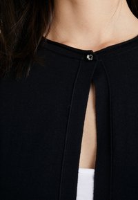 Schwarze Strickjacke mit glatter Textur, rundem Ausschnitt und einem Schlüsselöffnung, die mit einem kleinen Knopf gesichert ist.