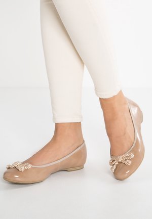 Beige ballet flats i lakeret læder med dekorativ snoet sløjfe, båret med hvide tætsiddende bukser på en lys baggrund.